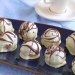 A plate of No-Bake Baileys Cheesecake Balls coated in white chocolate with a drizzle of caramel.